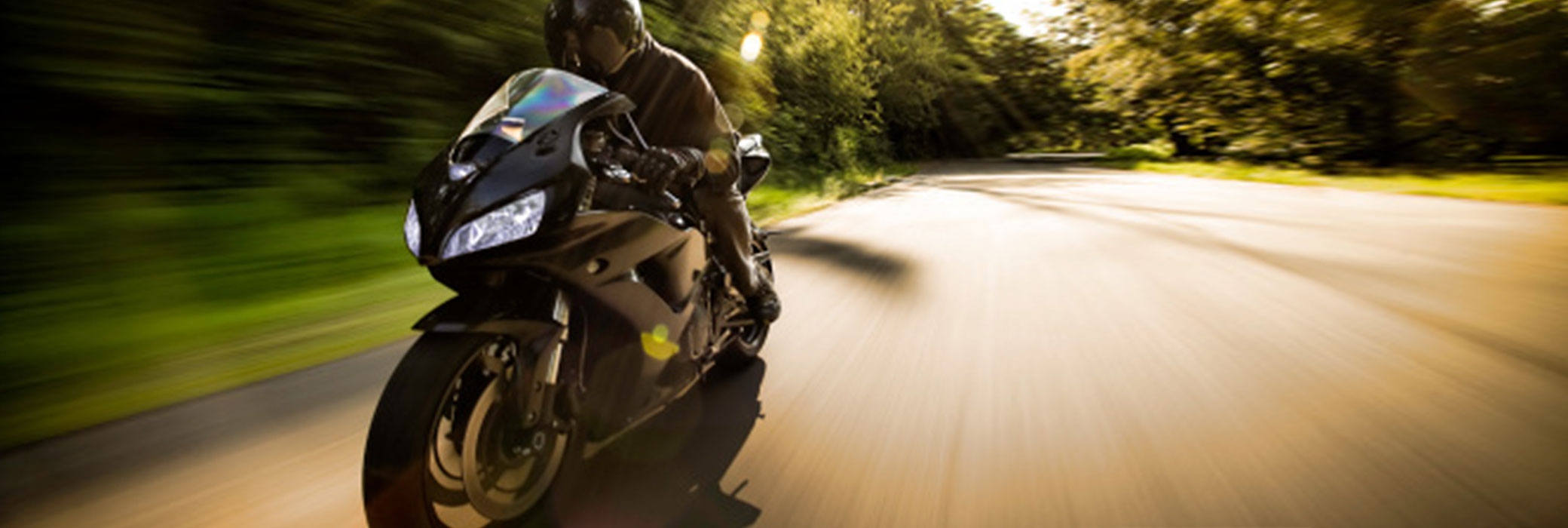Hombre vestido de negro que conduce una motocicleta negra por una carretera rural con árboles a los lados